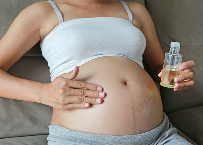 A heavily pregnant woman sitting on her coach rubbing citrus oil on her belly to prevent stretch marks