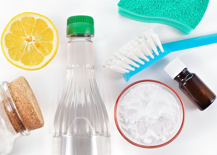A top-down view of different natural cleaning products such as a bottle of essential oil, lemon, vinegar, and baking soda