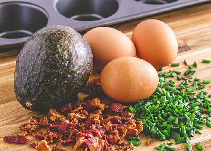 Ingredients for a Keto breakfast burrito prepped on a wooden board including fried bacon, spring onions, eggs, and an avocado