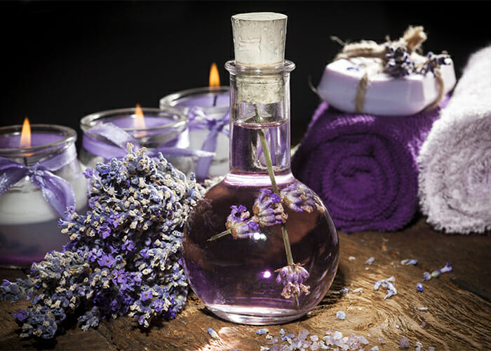 A glass potion bottle filled with homemade lavender essential oil next to a variety of lavender essential oil products such as candles and soaps.