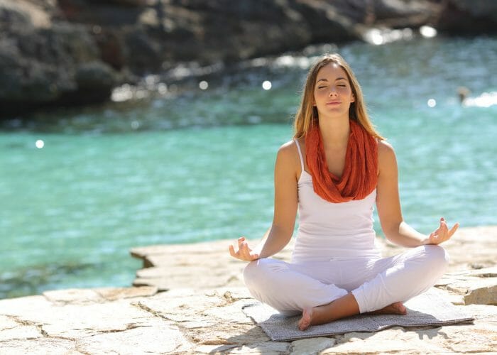 Woman wearing white clothes sitting by a river in a meditation pose doing breathing exercises