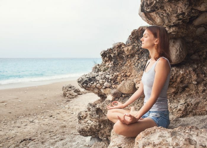 woman sat on a rock by the beach with eyes closed practicing breathing exercises