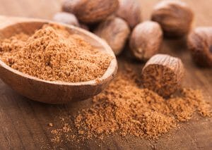 A wooden spoon filled with nutmeg powder next to fresh nutmeg
