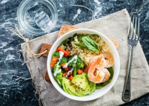 A healthy salad bowl with grains, vegetables, and shrimp, set on a wooden board on a blue marble table with a fork next to it