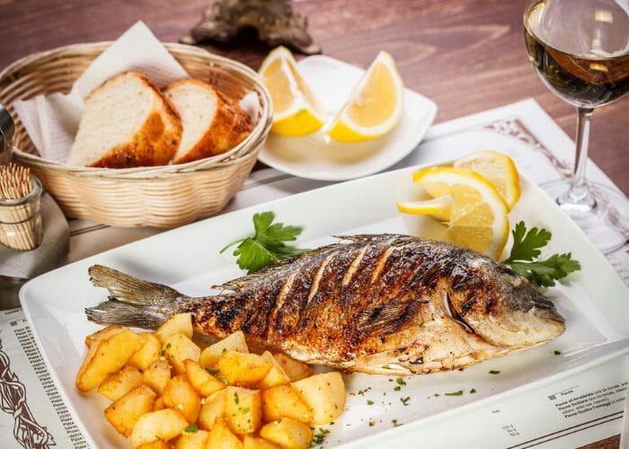 A dinner table with a basket of bread, glass of wine, and a whole grilled fish served on a white plate with a side of roasted potatoes and lemon wedges