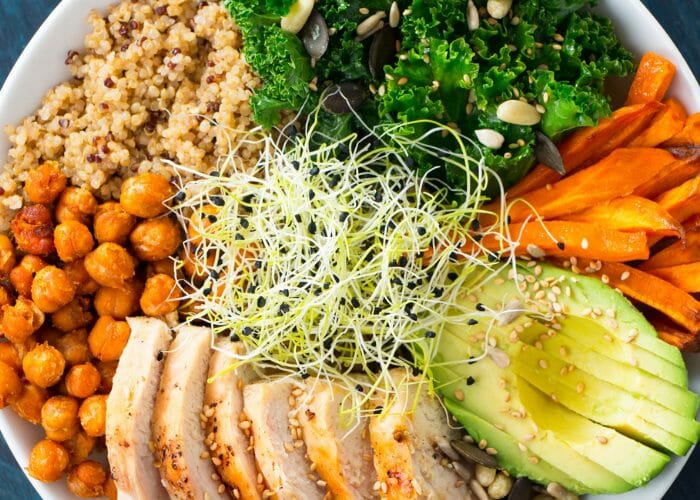 A semi-vegetarian salad bowl with quinoa, chickpeas, avocado, carrots, kale, seeds, alfalfa sprouts, and slices of grilled chicken breast