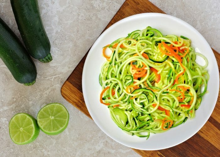 DIY spiralized zucchini and carrot noodles on a white dish, with two whole cucumbers and halved limes on the side