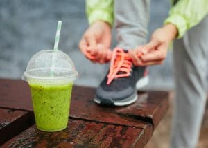 a cup of green smoothie on a wooden table with a woman tying her sports shoe shoelace in the background