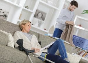 woman with leg in cast resting on a sofa with a carer doing chores