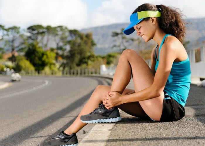 woman in running gear sitting on a roadside curb holding onto her ankle in pain