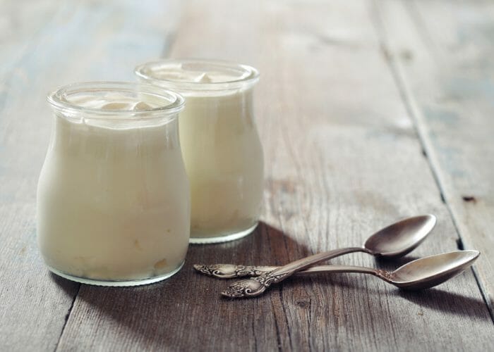 Two small, clear jars of homemade yogurt with two teaspoons next to them on a wooden table