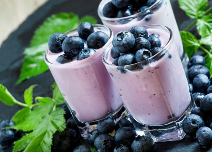 Three mini glasses filled with homemade blueberry yogurt on a dark tablecloth with blueberries and mint leaves around