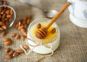 A small jar of homemade yogurt topped with honey and whole almonds, with a honey dipper on the top of the jar, and almonds spilled next to it