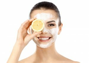 Woman in a face mask holding up half a lemon to her eye while smiling