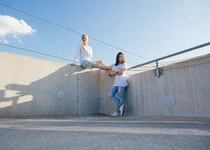 two women on a rooftop, one sitting on the edge of a wall and the other standing beside her friend