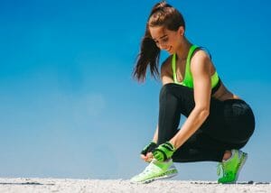 parkour woman dressed in neon green sports bra, black pants, green shoes, and green parkour gloves tying her shoelace