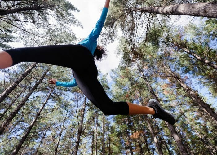 Bottom to top view of woman doing parkour training jumping across an obstacle in the woods surrounded by tall trees
