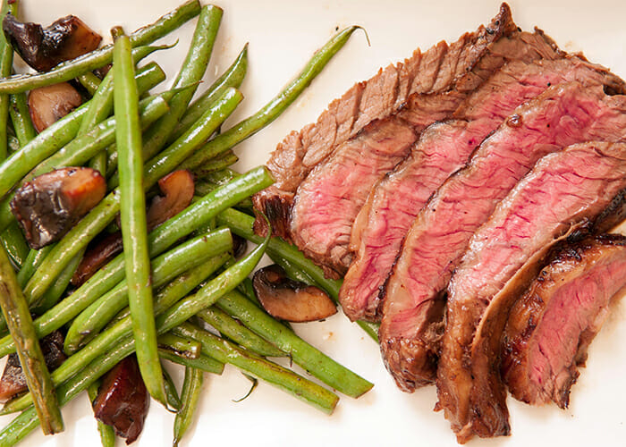 A plate of sliced medium-rare steak next to a side of green beans and mushrooms