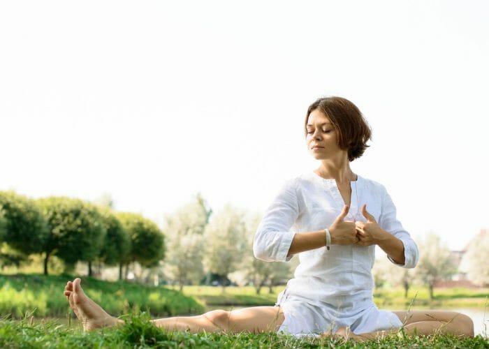 Woman in white Kundalini yoga clothes sitting on the grass in a park practicing a Kundalini yoga pose