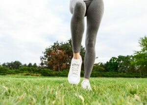 woman's legs with grey fitness leggings and white sneakers on green grass