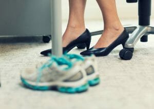 a professional woman seated at her desk in heels with running shoes set beside her