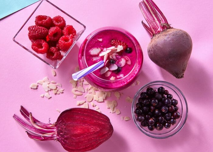 a glass of beet and berry juice topped with almonds, a cup filled with raspberries and blueberries and fresh beets around it 