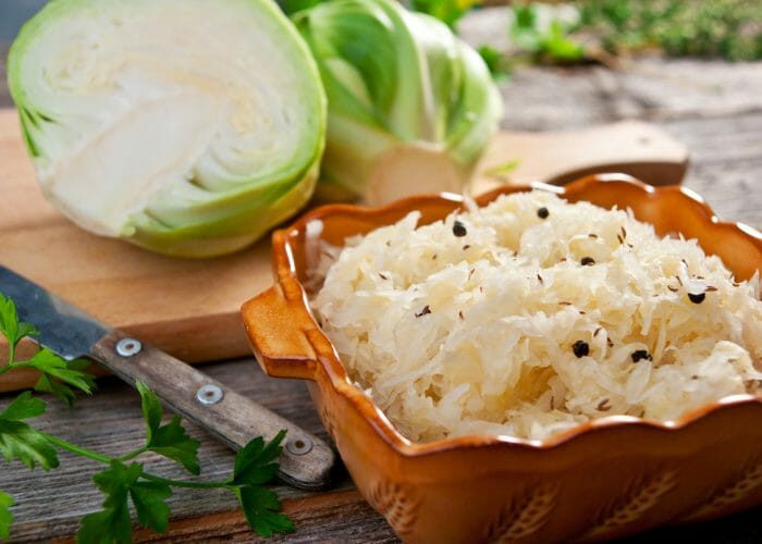 Sauerkraut in a brown ceramic bowl with cabbage halves in the background on a wooden chopping board
