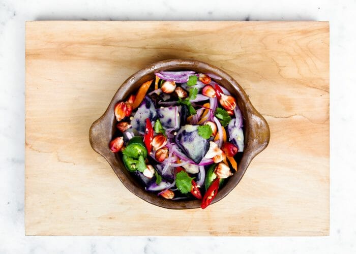 Fresh colorful purple, red, and green vegetables in a dutch oven, on top of a wooden board, set against a light marble background