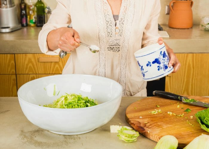 Woman making her own sauerkraut at home pouring sea salt into a large white bowl with sliced cabbage inside