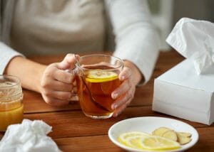woman with the flu sat at a table holding a glass of lemon and ginger tea with tissues scattered around