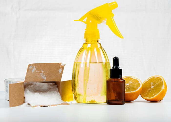 Different types of natural cleaners without chemicals such as a bottle of citrus essential oil, a spray bottle of vinegar, and a box of bicarbonate soda