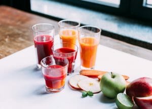 Tall gasses of fresh fruit juice, with whole fresh fruits and sliced fruits beside it on a white table