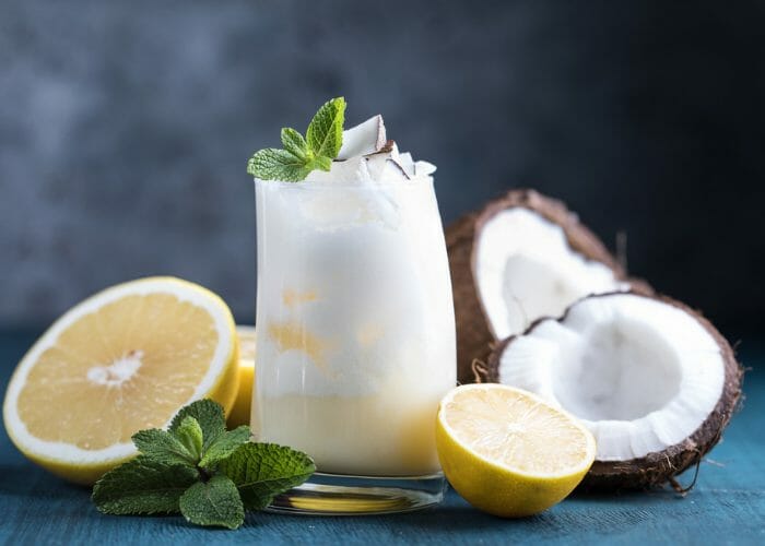 a glass of coconut smoothie with fresh coconut, lemons and mint leaves around it.