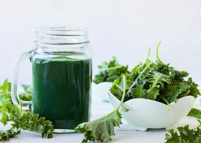 a green mason jar cup of green smoothie with a bowl of kale leaves next to it and kale leaves scattered around