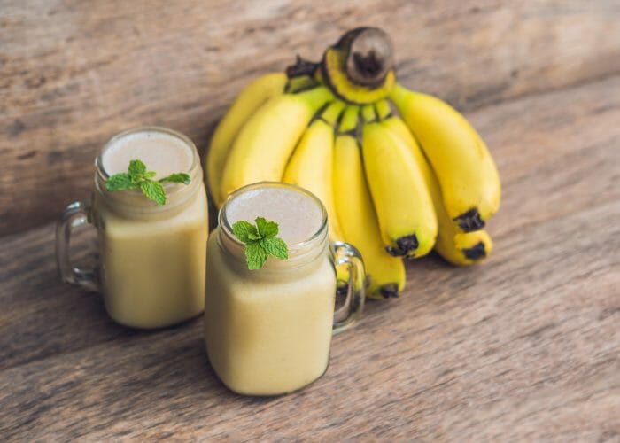 two mason jar glasses of banana oat smoothies, garnished with sprigs of mint, and a bunch of bananas set behind them on a wooden table