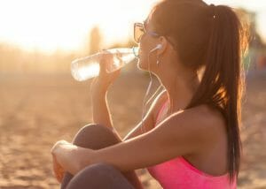 woman with earphones and sunglasses drinking water from her bottle after her run on the beach