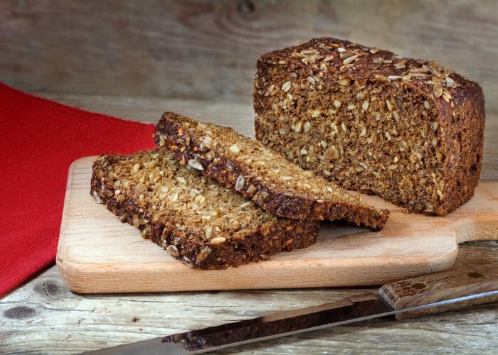 Loaf of protein bread with whole grains and seeds sliced on a wooden board with a red tablecloth in the corner