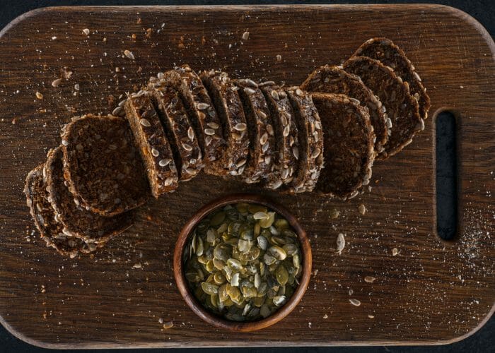 Sliced loaf of dark protein bread with a small bowl of pumpkin seeds on a dark wooden board
