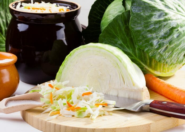 Sliced cabbage and a half cabbage on a chopping board with knife, with pots of sauerkraut in the background