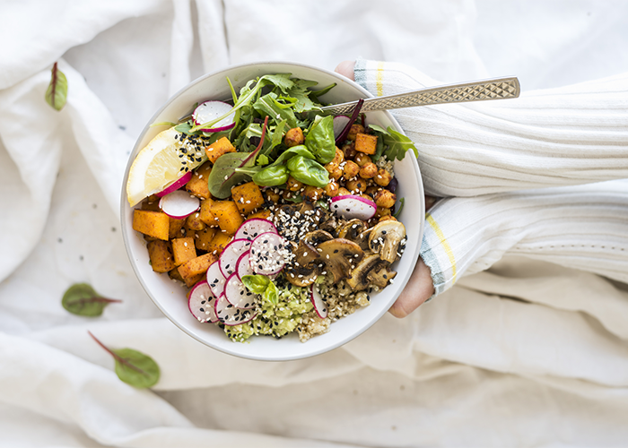 Woman in sweater holding up a vegan Buddha bowl filled with greens, pumpkin, beets, mushrooms, seeds, a wedge of lemon