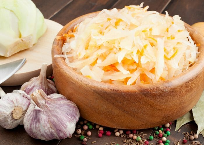 Large wooden bowl filled with sauerkraut, with ingredients like cabbage and garlic scattered around it on a wooden table