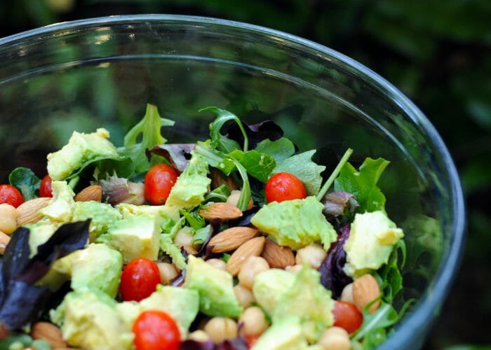 A mixed salad of green vegetables, cherry tomatoes, avocado, almonds, and chickpeas in a glass salad bowl