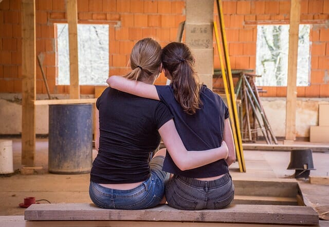 Two femasle friends sat side by side with their backs to the camera. they ahve their arms around one another to help support and show their friendship. They are in what looks like a garage or work room.